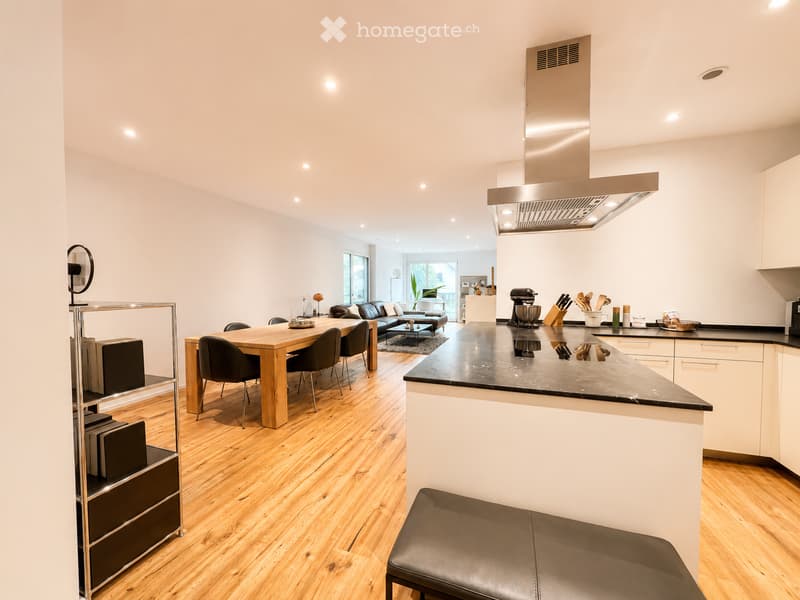 Open-plan kitchen and dining area with light wooden flooring, white walls, and modern fixtures. Features a central dining table with chairs, a sofa in the background, and a kitchen island with a black countertop and various appliances.