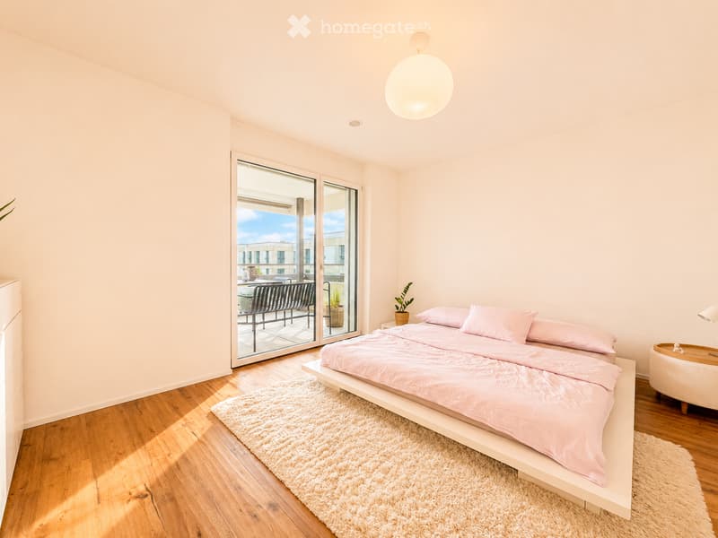 Modern bedroom with light pink bedding, a white nightstand, a round table lamp, a fluffy white rug, and a balcony with outdoor seating visible through large glass doors.