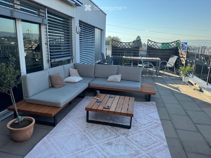 Balcony with a grey sectional sofa, wooden coffee table, dining table with chairs, and potted plants.