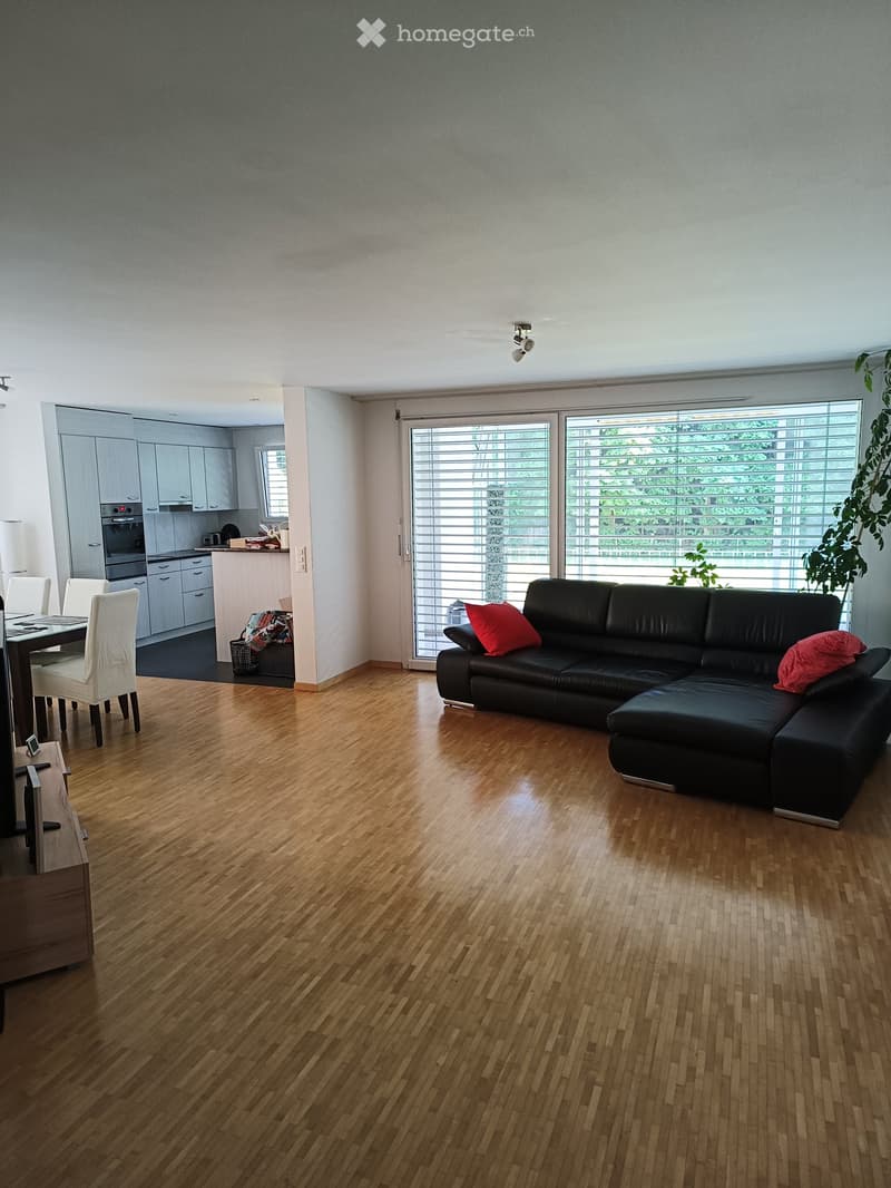 Modern living room with hardwood flooring, large windows, a black sectional sofa with red cushions, a dining table with chairs, and an adjacent kitchen area with cabinets and an oven.