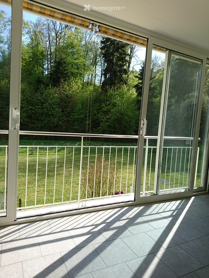 Spacious balcony with large glass doors, white metal railing, and a view of a green lawn and trees.