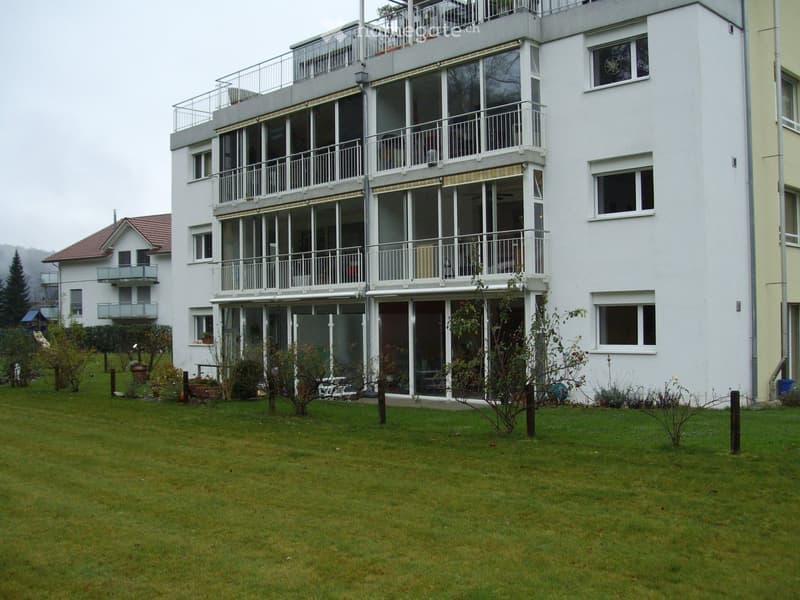 Modern multi-story apartment building with large windows and balconies, surrounded by a well-maintained lawn and small garden area.