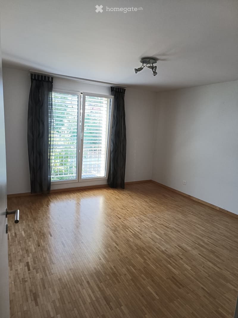 Empty room with wooden flooring, large windows with dark curtains, and a ceiling light fixture.