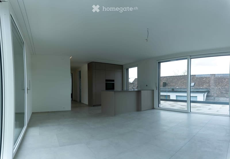 Empty room with tiled floor, large windows, and a small kitchen area with cabinets and a countertop.