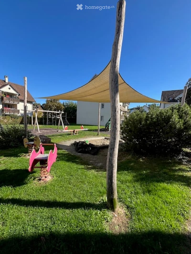A sunny outdoor play area with a grassy lawn, a large canopy, and colorful play equipment including a pink seesaw and a swing set.