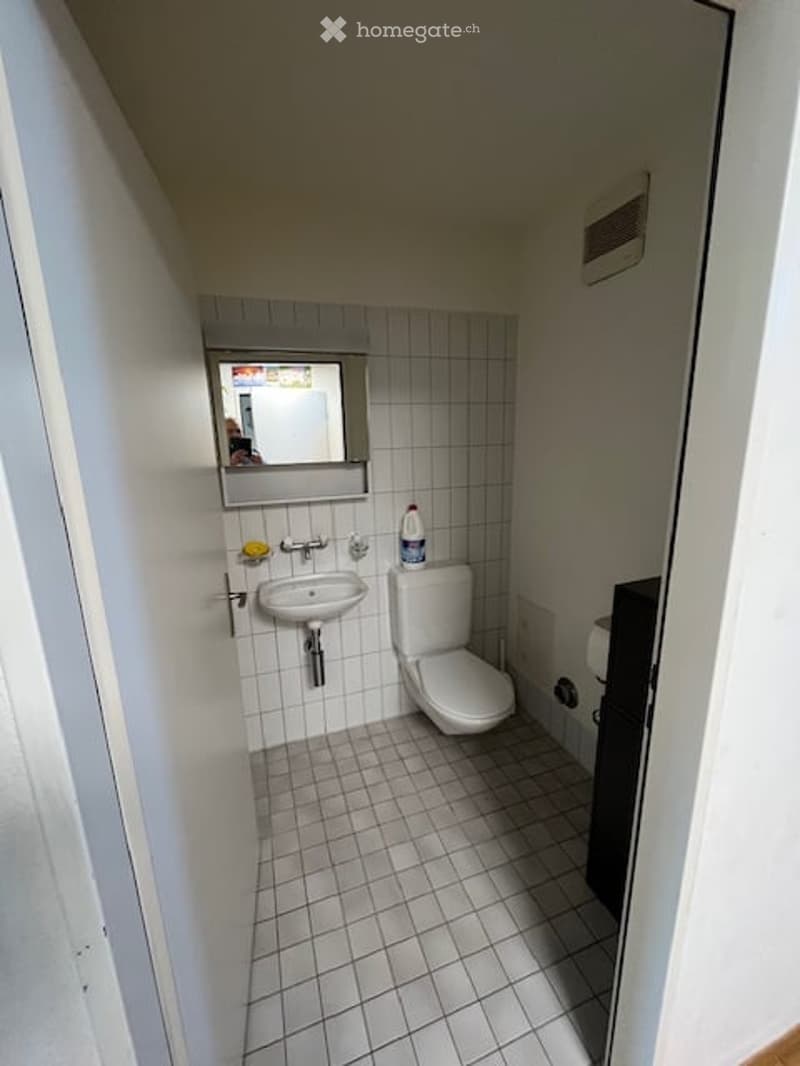 Small bathroom with white tiled walls and floor, a sink, toilet, and mirror above the sink.