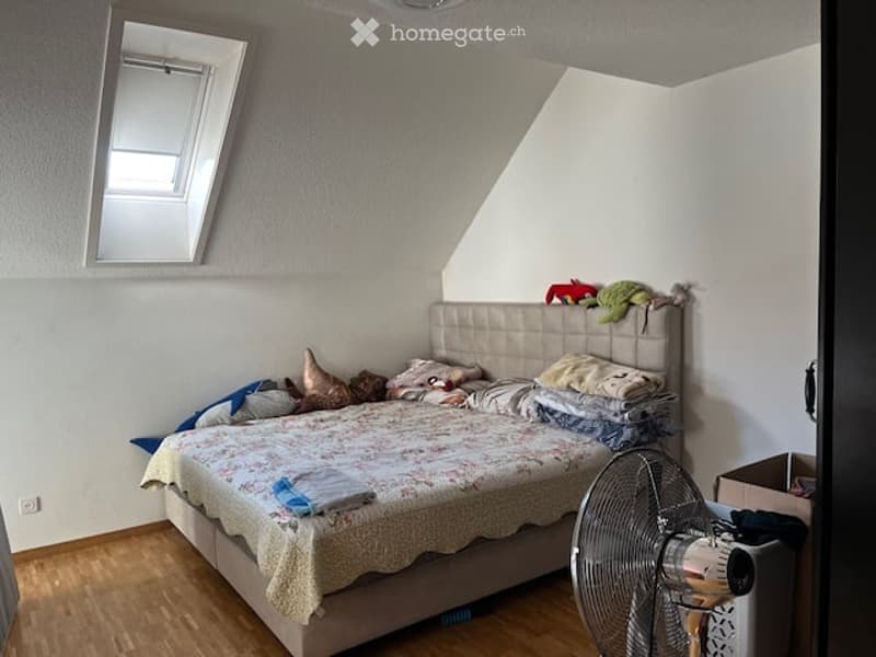 Bedroom with a large bed, pillows, blankets, and a fan. The room has wooden flooring and white walls.