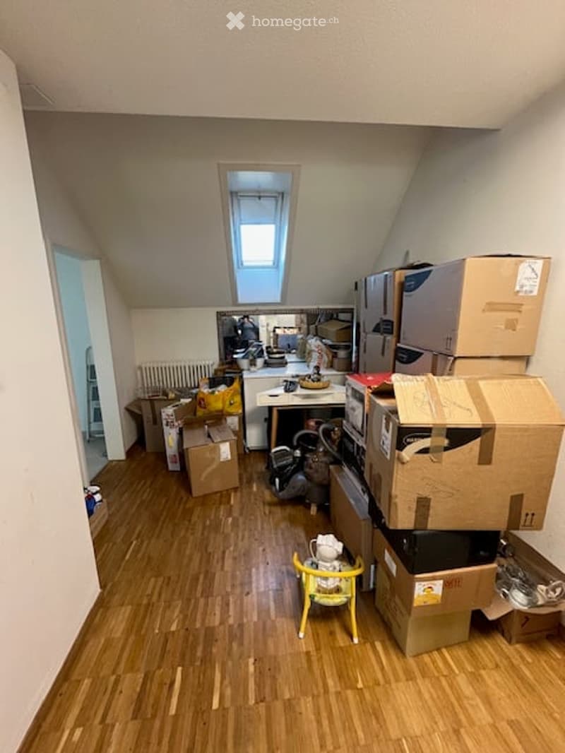 An attic space with wooden flooring, slanted ceilings, and a skylight window. The room is empty except for a few boxes and a small yellow chair, indicating it is unfurnished.