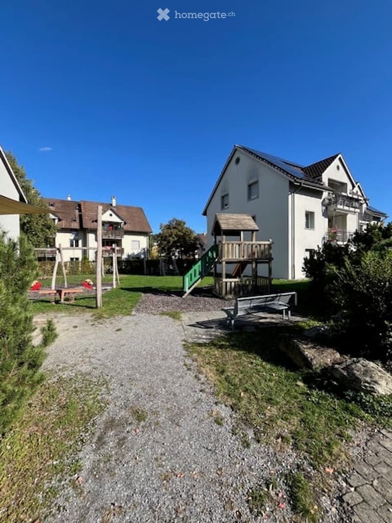 A well-maintained outdoor play area with a wooden play structure, swings, a bench, and surrounding greenery.
