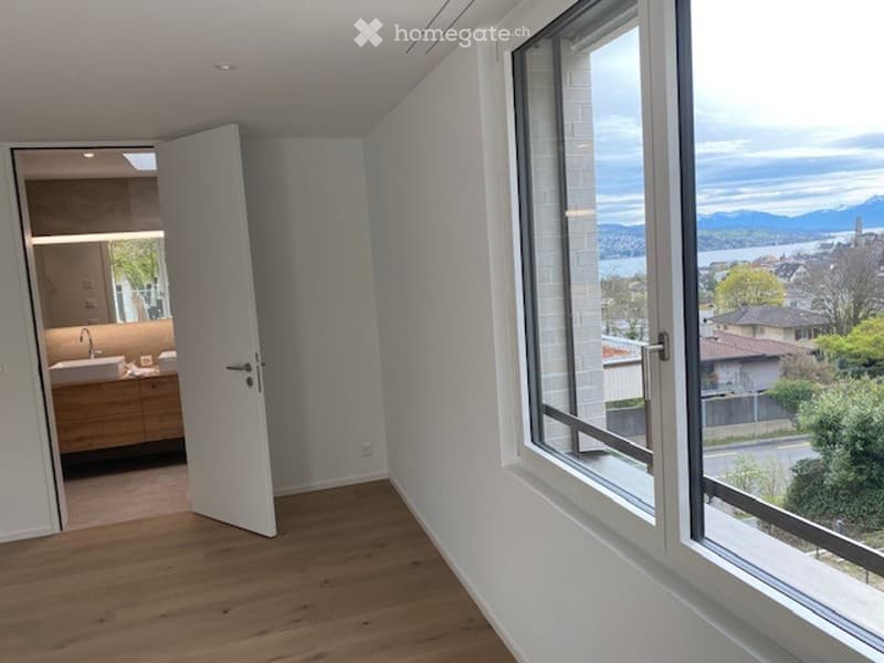 A room with a white door and white walls, a wooden floor, and a window that provides a view of a lake and mountains.
