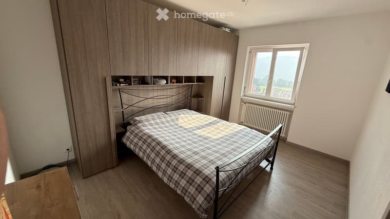 Modern bedroom with a wooden floor, large window with a scenic view, a metal-framed bed with a plaid duvet, a tall wardrobe with shelves, and a nightstand.