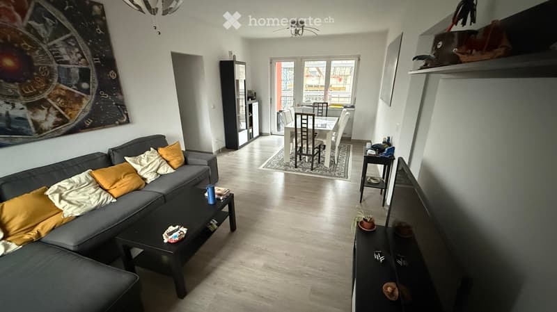 Open-plan living and dining area with a grey sectional sofa, coffee table, dining table with chairs, and a large window leading to a balcony.