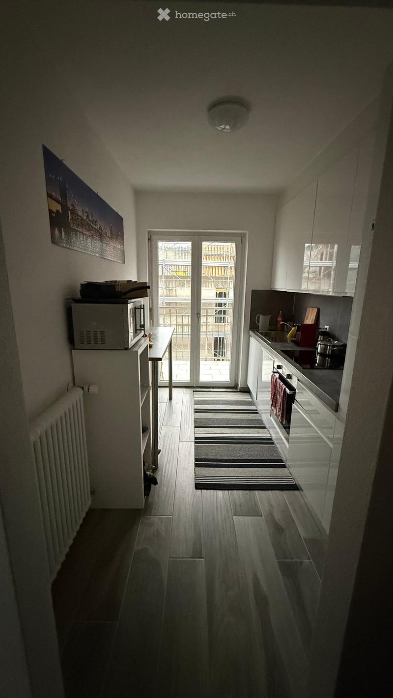 Modern kitchen with white cabinets, a microwave, oven, sink, and a small table near the entrance. The floor is wooden, and there is a striped rug in front of the door leading to a balcony.