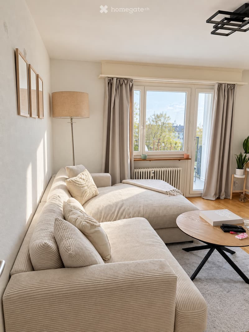 Modern living room with a sofa, coffee table, floor lamp, and large windows offering a view of trees and buildings.