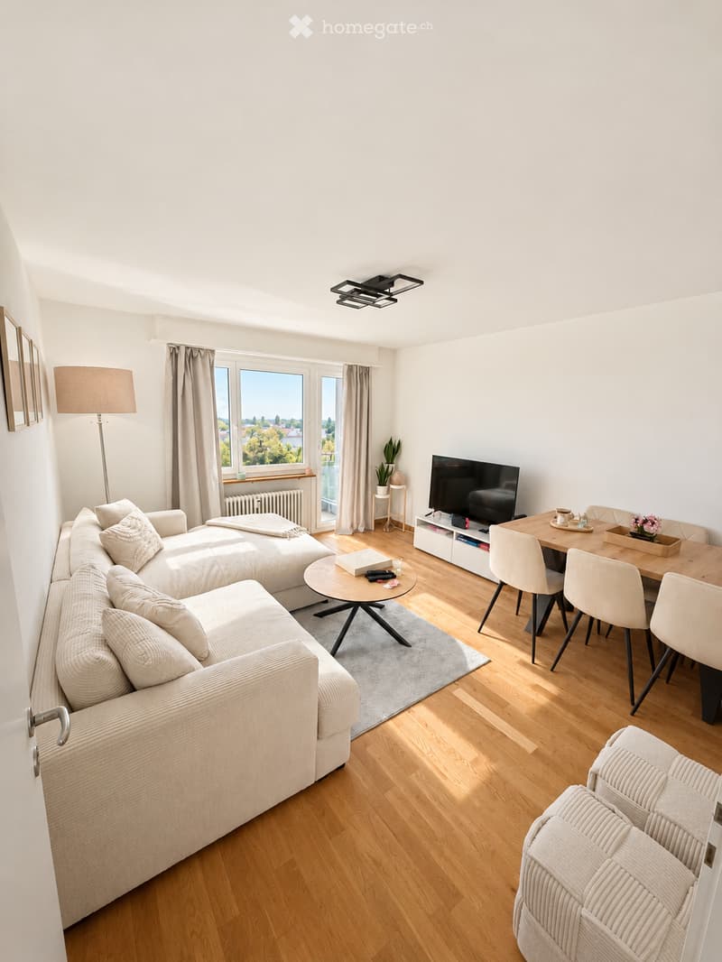 Modern living room with a large window offering a view, featuring a sectional sofa, coffee table, dining table with chairs, TV, floor lamp, and a small plant on a side table.