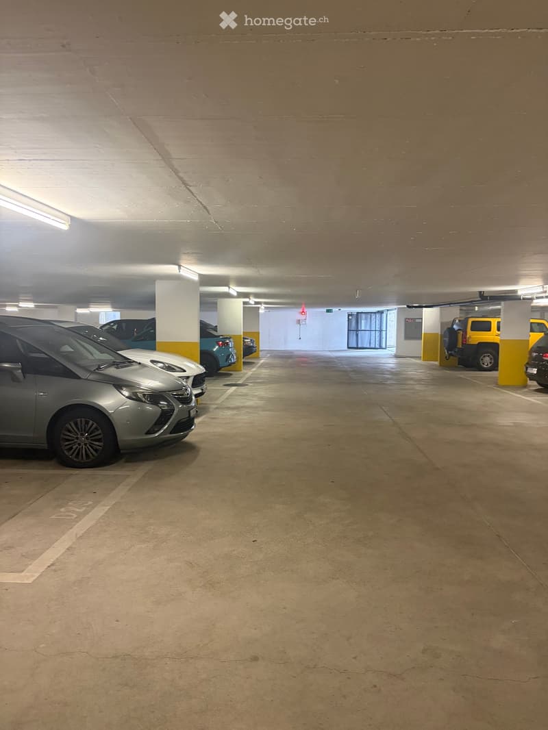Underground parking area with several parked cars, concrete pillars, and fluorescent lighting.