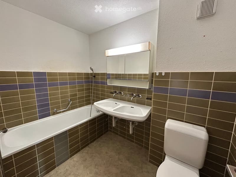 Modern bathroom with a bathtub, double sink, toilet, and a mirrored cabinet with built-in lighting. The walls are tiled in a brown and gray pattern, and the floor is covered with beige tiles.