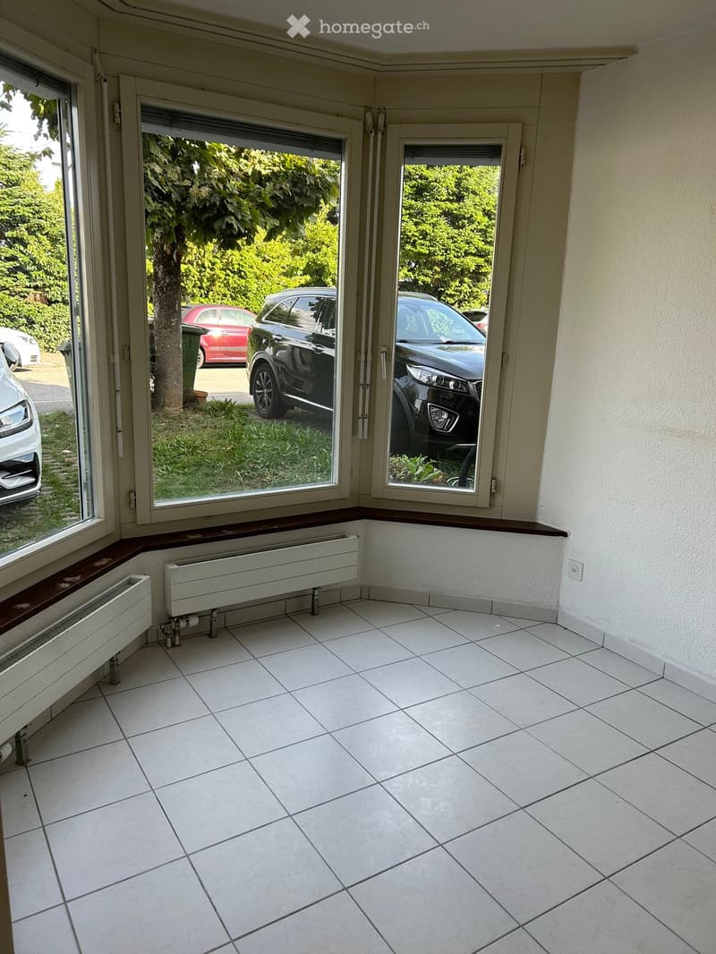 Empty room with white tiled flooring, large windows, and radiators along the walls. The room is unfurnished and has a view of parked cars and greenery outside.