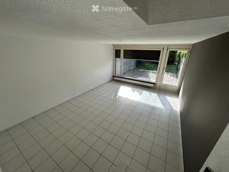 Empty room with tiled floor, white walls, large sliding glass doors leading to a wooden deck and garden area.