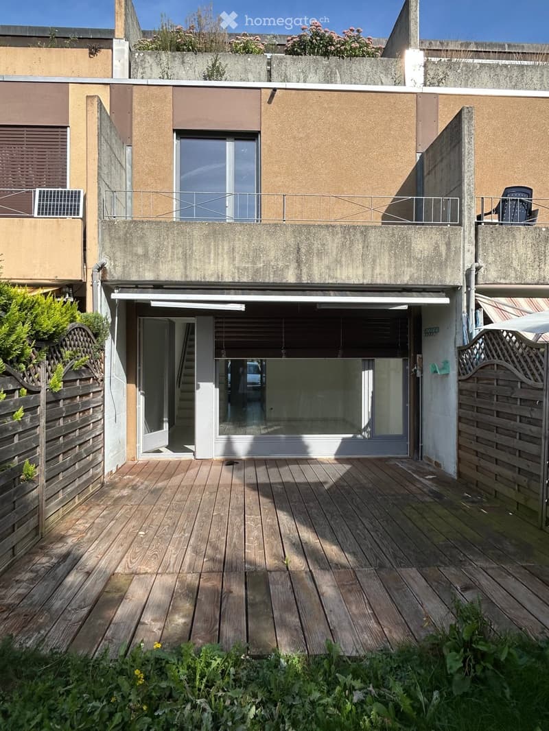 Wooden deck area with plants and a small garden, adjacent to a modern building with a balcony and garage entrance.