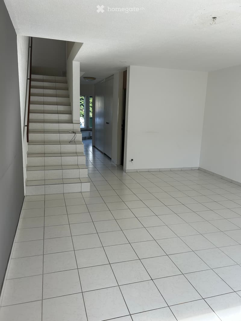 Empty hallway with tiled flooring, white walls, and a staircase leading upwards. Natural light filters in from a window at the far end.