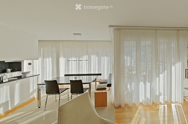 Bright, modern living and dining area with large windows covered by sheer curtains, a sleek black dining table with chairs, a wooden bench, and a white sofa. The kitchen area features a black countertop with a cup and bowl.