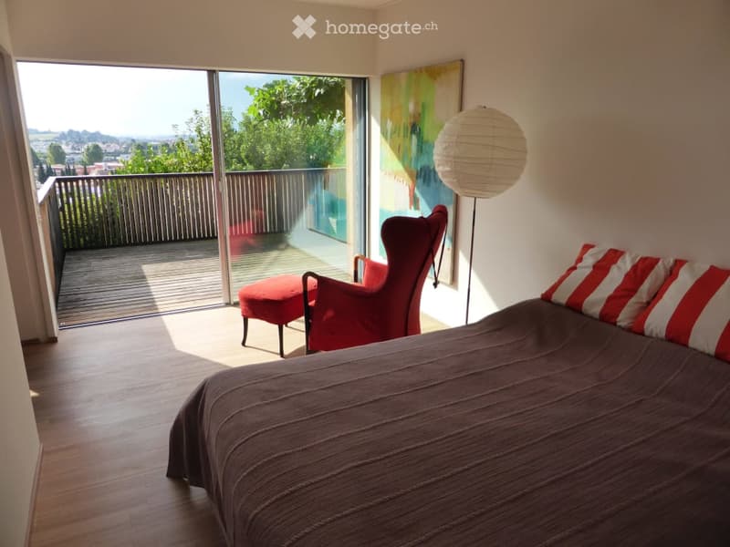 Bedroom. Modern bedroom with wooden flooring, large window offering a view, red armchair, red ottoman, and a floor lamp with a white shade.