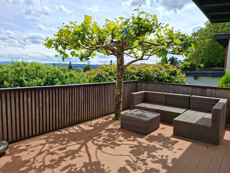 Balcony / Terrace. Wooden deck with a wicker sofa and table, surrounded by greenery and a tree, with a scenic view of the landscape.