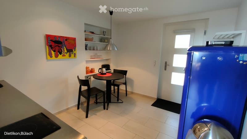 Furnished Room. A small, modern kitchen area with a round dining table, four chairs, a blue refrigerator, shelves with cups and a teapot, and a colorful painting on the wall.