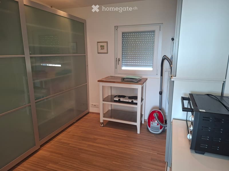 Furnished Room. A small, furnished room with wooden flooring, a table with an induction cooktop, a vacuum cleaner, and a refrigerator. There is a large glass door and a window with blinds.