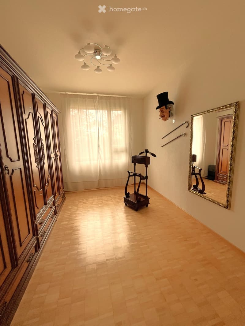 Empty hallway with wooden flooring, large window with white curtains, ornate wardrobe, mirror, and decorative items including a small table and a walking stick.