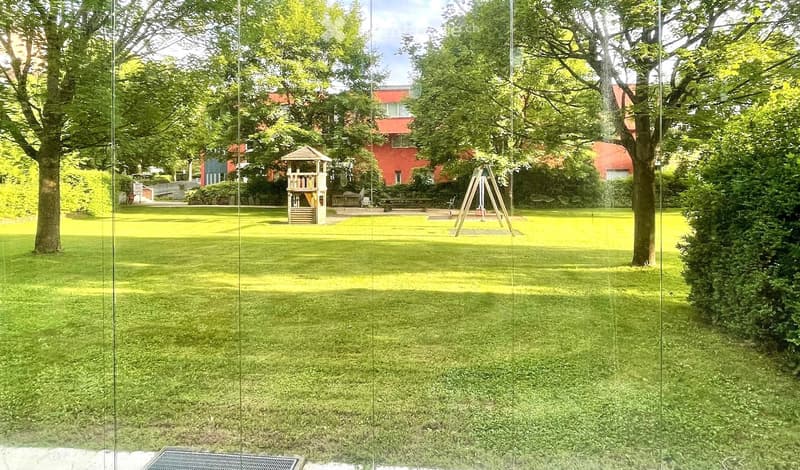 Well-maintained lawn with trees, a small playhouse, and a swing set in the background, with residential buildings visible beyond the trees.
