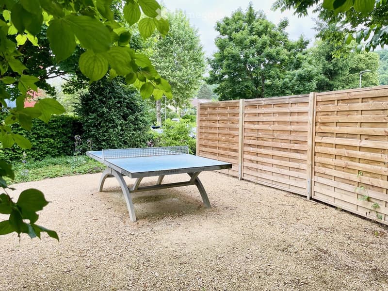 Outdoor area with a table tennis table, wooden privacy screens, gravel ground, and lush greenery.