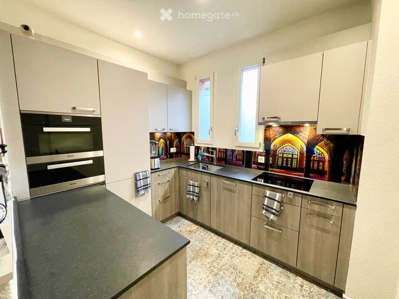 Modern kitchen with light wood cabinets, stainless steel appliances, and a decorative backsplash featuring stained-glass designs.