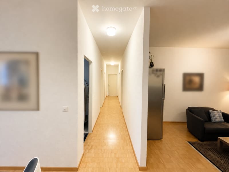 A modern hallway with wooden flooring, white walls, and recessed lighting. Visible furniture includes a sofa, a small table, a chair, and a refrigerator.
