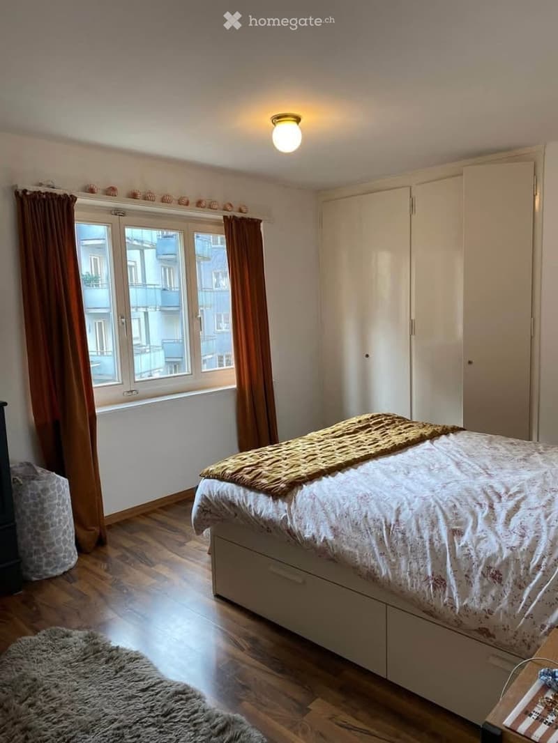 Cozy bedroom with wooden flooring, large window with brown curtains, a neatly made bed with a patterned duvet and a textured throw, a built-in wardrobe, and a soft area rug near the bed.