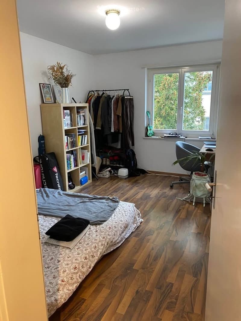 A cozy bedroom with wooden flooring, a bed with a patterned duvet, a bookshelf filled with books, a clothing rack, a guitar on a stand, a desk with a chair, and a plant near the window.
