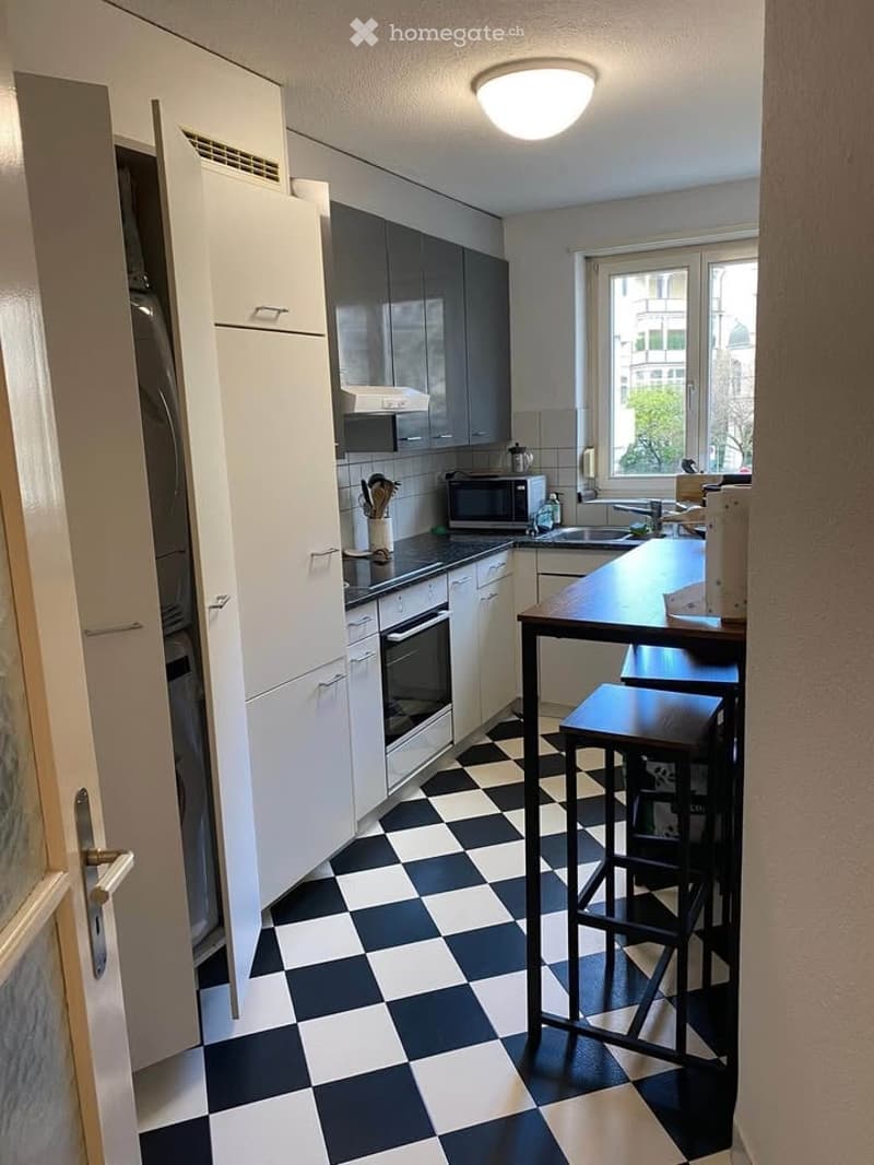 Modern kitchen with white cabinets, black countertops, and a checkered black and white floor. Features include a refrigerator, oven, microwave, sink, and a small dining area with a table and stools.