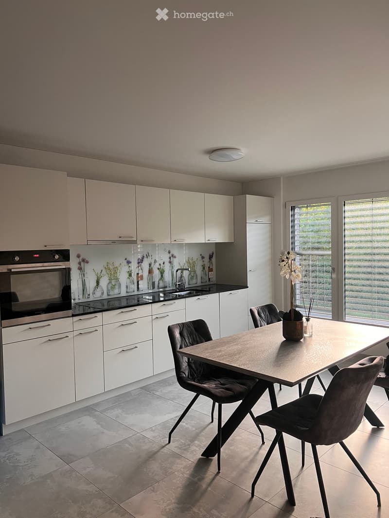 Modern kitchen with white cabinets, a central dining table with four chairs, and a built-in oven. The space features a sleek design with a tiled floor and large windows allowing natural light.