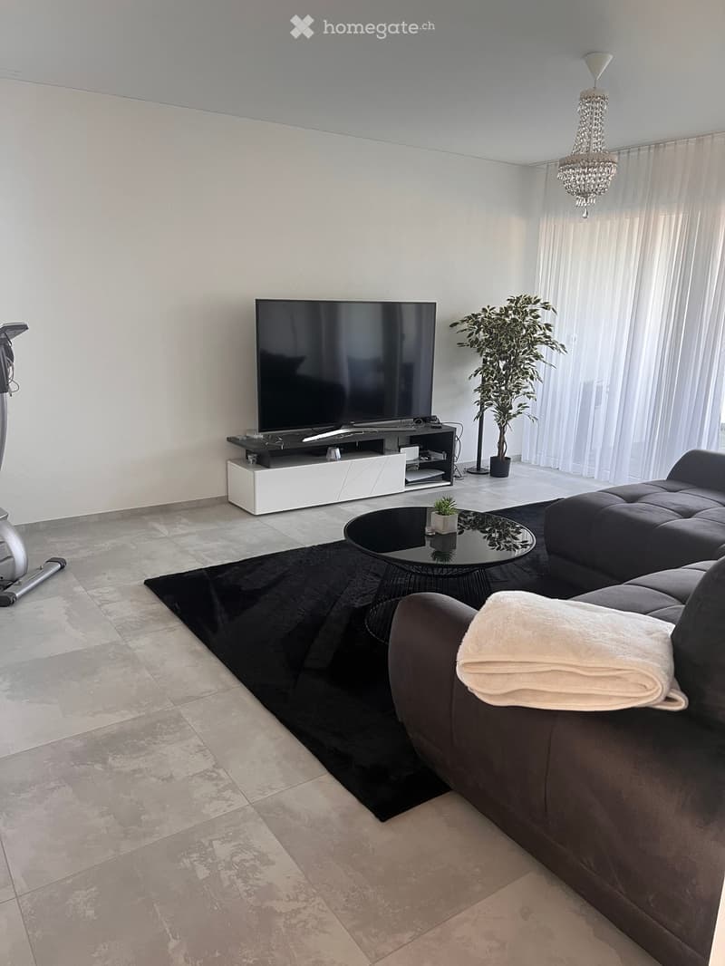 Modern living room with a dark grey sofa, a black coffee table, a large flat-screen TV on a white stand, a chandelier, and a potted plant near large windows with sheer curtains.