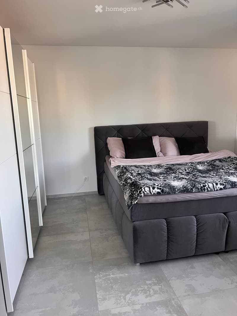 Modern bedroom with a large bed featuring a tufted headboard, multiple pillows, and a fur throw. The room has light grey walls and a concrete floor.