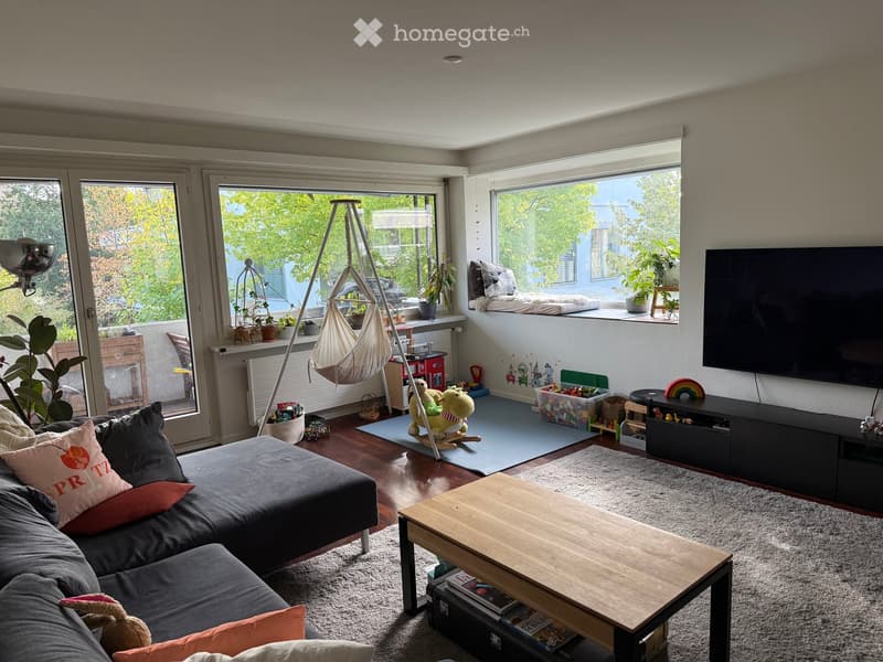 Living room with large windows, grey sectional sofa, wooden coffee table, TV stand, hanging chair, toys, and plants.