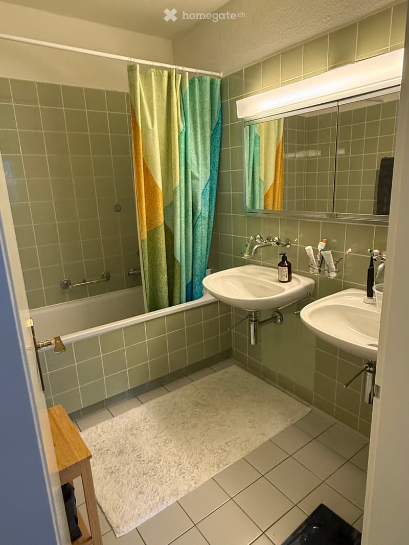 Bathroom with green tiled walls, bathtub with shower curtain, two sinks, mirror, and a small wooden shelf.