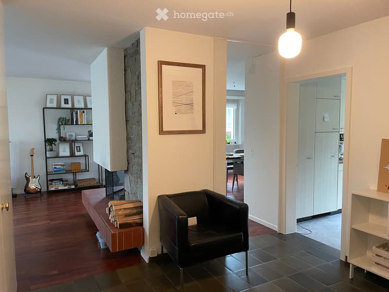 A modern living room with a black leather chair, wooden bench, shelf with books and a guitar, and a picture frame on the wall. The room has wooden flooring and a view into a kitchen area.