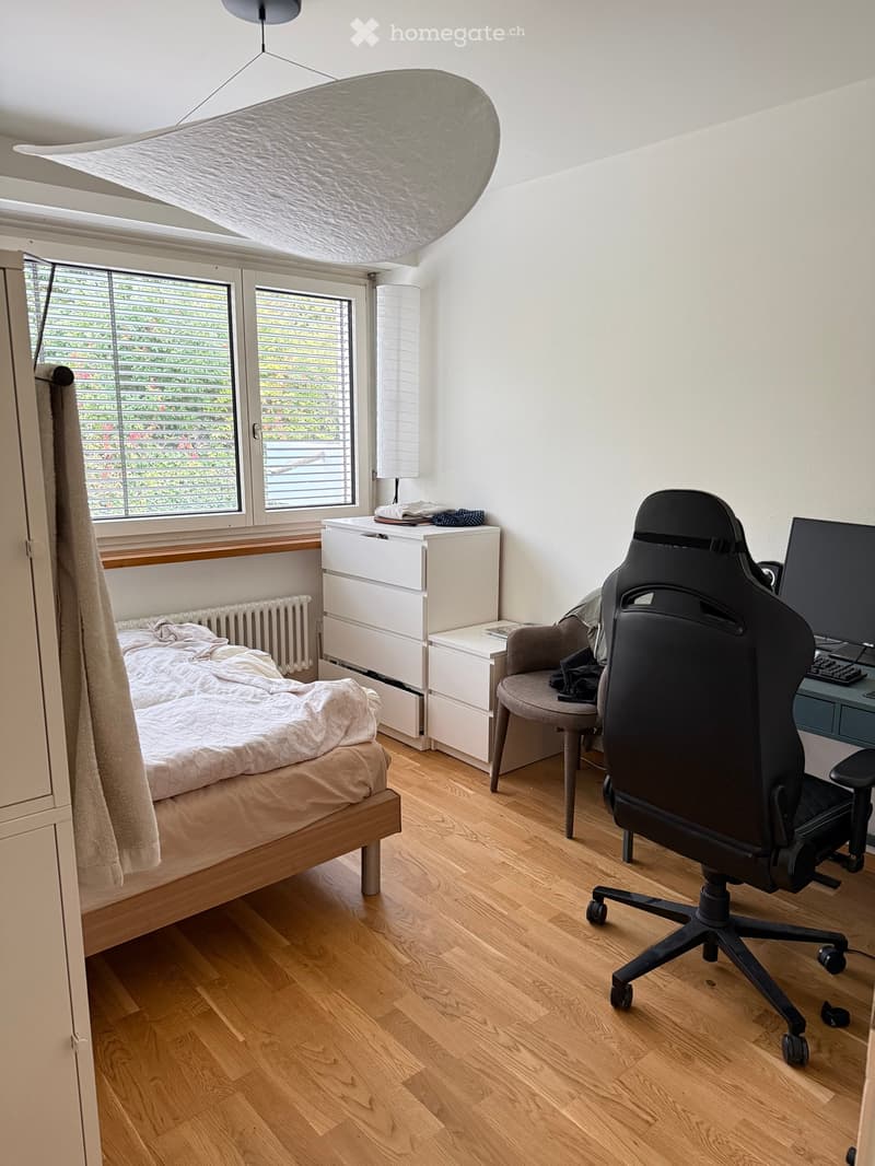 Bright bedroom with wooden flooring, large windows, a bed with white bedding, a desk with a computer setup, and a comfortable chair.