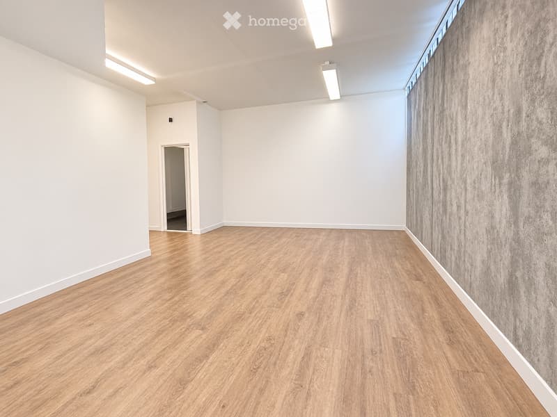 Empty room with light wooden flooring, white walls, and a grey textured accent wall. A doorway leads to another space.