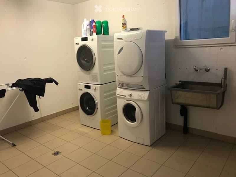 Laundry room with stacked washing machines and dryer, sink, and cleaning supplies on top of the machines.