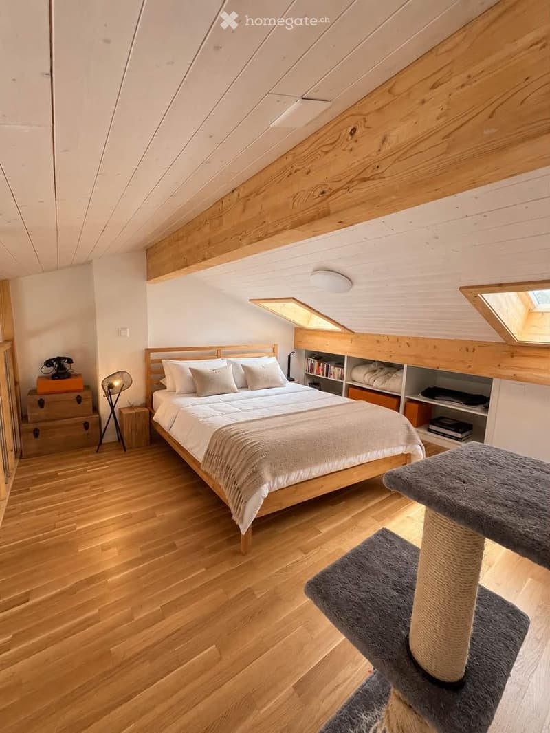 Cozy attic bedroom with wooden floors, slanted wooden ceiling, skylights, a large bed with white bedding, a cat tree, a nightstand with a lamp, and a vintage telephone on a chest.