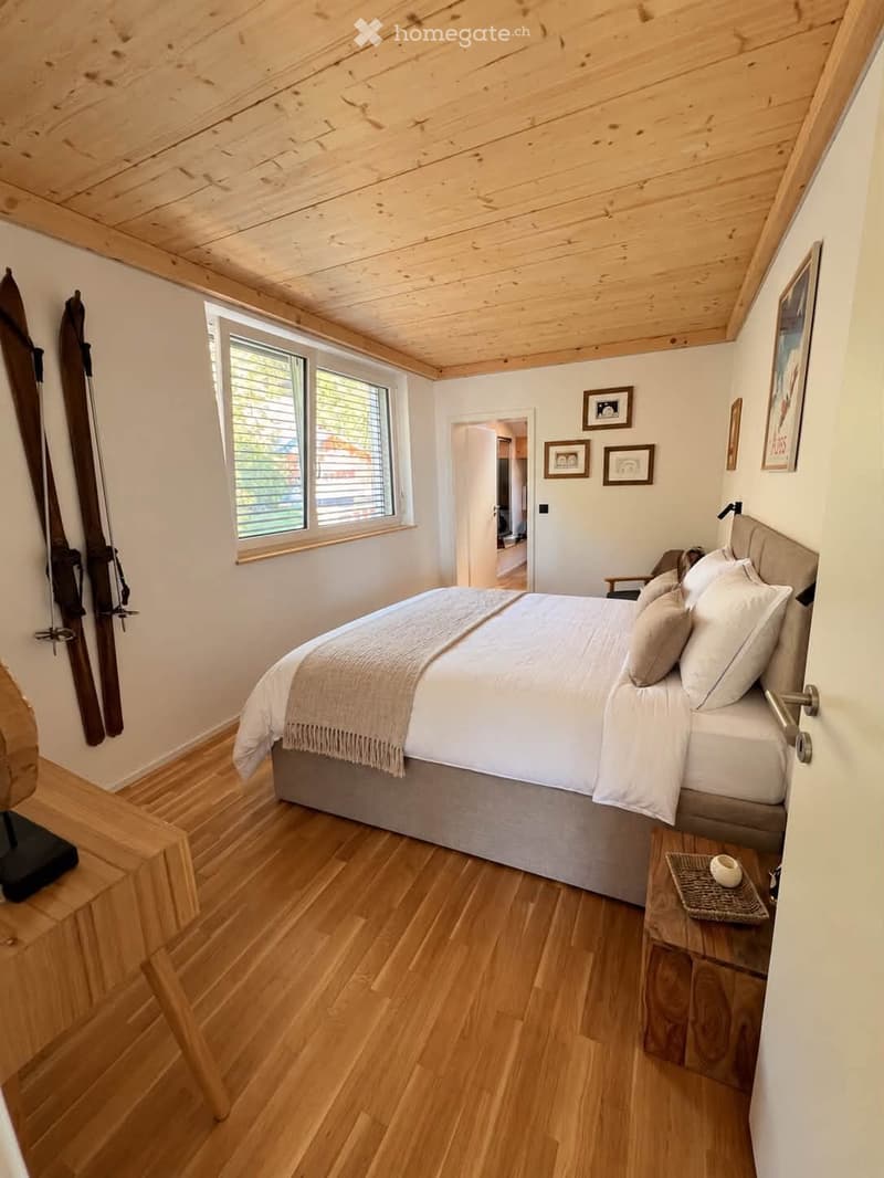 Cozy bedroom with wooden floors, slanted wooden ceiling, large window, bed with white linens, nightstand, and ski leaning against wall.