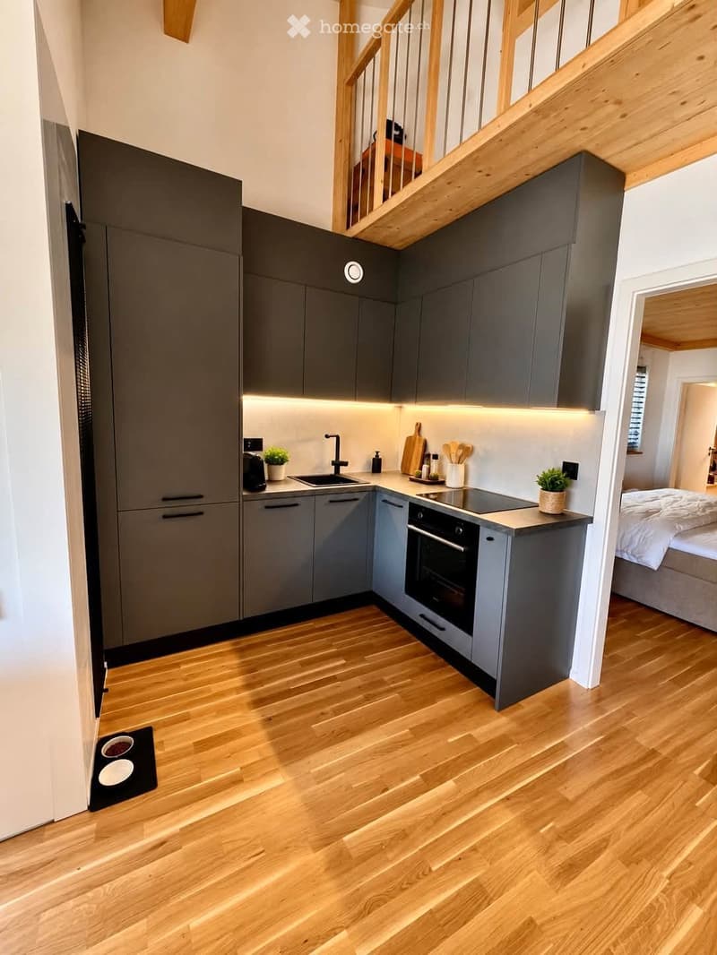 Modern kitchen with grey cabinetry, wooden flooring, and built-in appliances. A small bed is visible in the adjacent room.